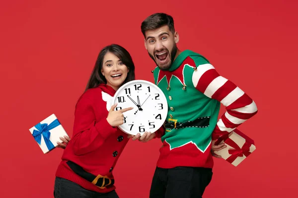 Glada par i roliga jul tröjor med vit stor klocka och lådor med gåvor över röd bakgrund. — Stockfoto
