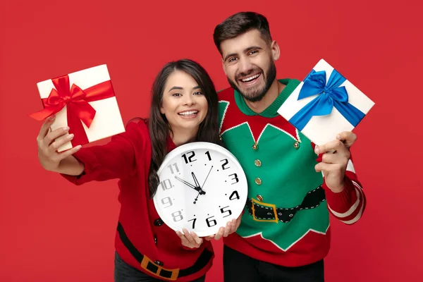 Sorrindo casal em camisolas engraçadas de Natal segurando branco grande relógio e caixas com presentes sobre fundo vermelho. — Fotografia de Stock