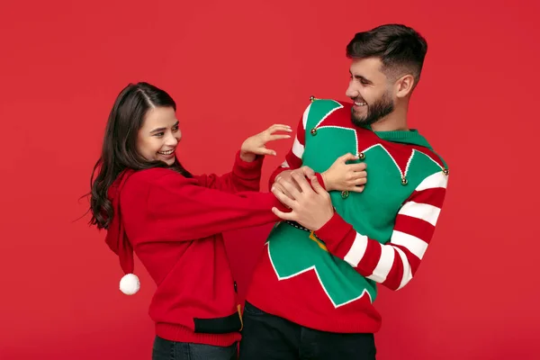 Casal positivo vestido com camisolas engraçadas de Natal. Mulher cócegas seu namorado sobre fundo vermelho. — Fotografia de Stock