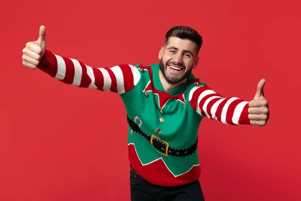 Alegre hipster homem vestido com engraçado Natal suéter mostrando polegares para cima sobre vermelho fundo. — Fotografia de Stock