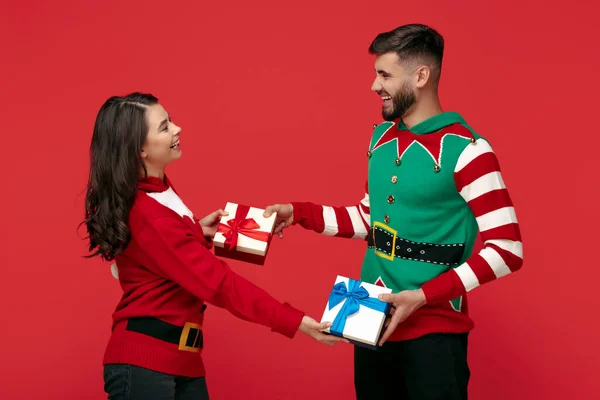 Encantador casal em camisolas engraçadas de Natal segurando caixas de presente sorrindo sobre fundo vermelho. — Fotografia de Stock