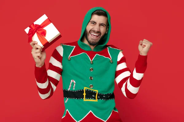 Homem hipster feliz vestido com camisola de Natal engraçado segurando caixa de presente e celebrar sobre fundo vermelho. — Fotografia de Stock