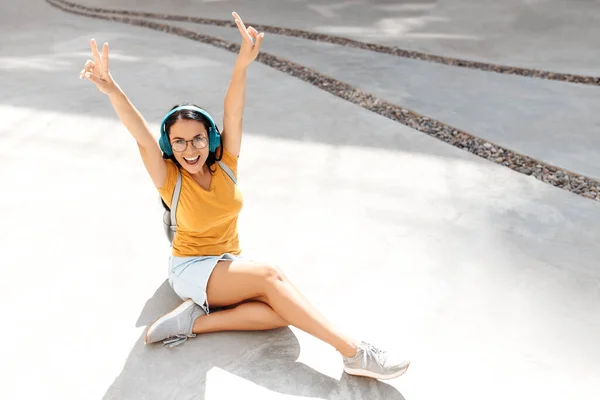 Aufgeregte brünette junge Studentin hört Musik über Kopfhörer, sitzt auf Zementboden vor rostiger Wand. — Stockfoto