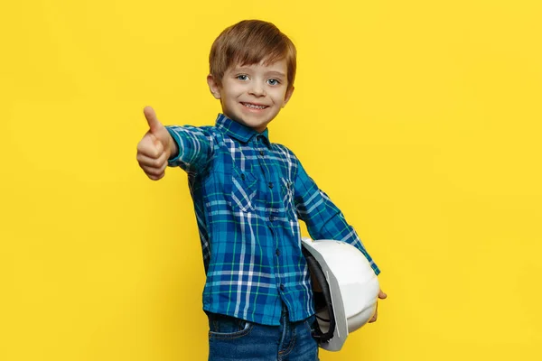 戴着保护头盔的积极的小男孩工程师或建筑师，在黄色的背景上竖起大拇指. — 图库照片