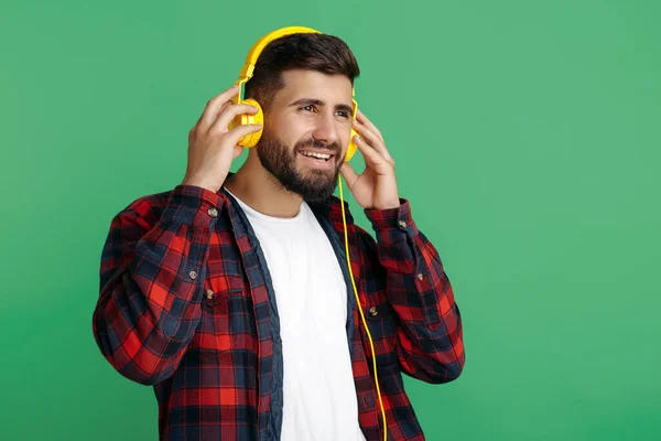 Bello barbuto hipster giovane in camicia a quadri ascoltare musica in cuffia con gli occhi chiusi su sfondo verde. — Foto Stock