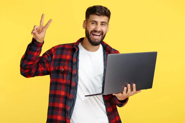 Opgewonden hipster jongeman toont vrede gebaar op notebook of computer over gele achtergrond. — Stockfoto