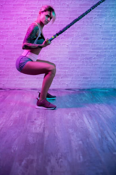 Beautiful sporty young blonde woman workout with stretch bands in gym in neon lights. — Stock Photo, Image