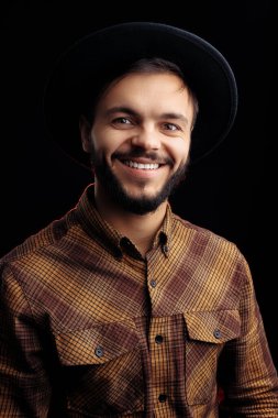 Kahverengi gömlekli yakışıklı, sakallı hippi siyah arka planda gülümsüyor..