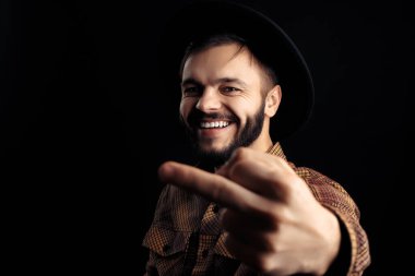 Kahverengi gömlekli ve siyah şapkalı gülümseyen genç sakallı hippi siyah arka planda orta parmağını gösteriyor..