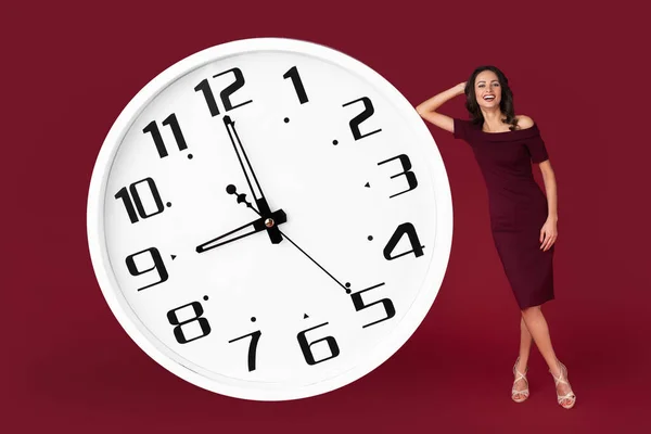 Fröhlich elegante junge moderne Geschäftsfrau im kastanienbraunen Kleid lehnt an einer riesigen weißen Uhr vor rotem Hintergrund. — Stockfoto