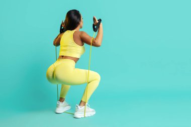 Çekici, genç, esmer, sarı spor kıyafetli kadın. Mavi arka planda bantla çömelme hareketleri yapıyor..