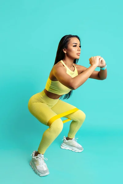 Sportovní mladá brunetka žena ve žluté sportovní oblečení dělá dřepy s strečovou páskou přes modré pozadí. — Stock fotografie