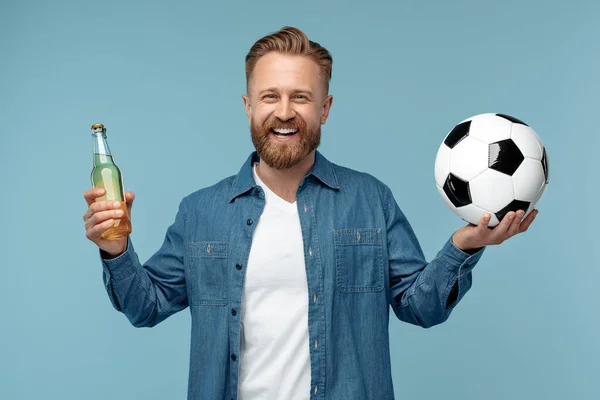 在演播室里，快乐的金发嬉皮士男人拿着经典足球和一瓶啤酒，背景是蓝色的. — 图库照片