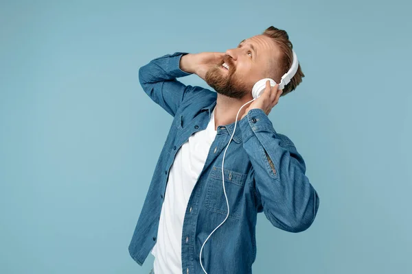 Stijlvolle bebaarde blonde hipster man in jeans shirt met koptelefoon over blauwe achtergrond. — Stockfoto