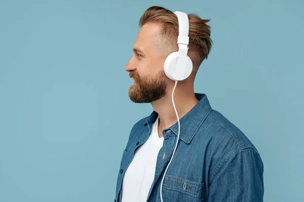 Portret van een bebaarde blonde hippe man in een jeans shirt met een koptelefoon en wegkijkend over een blauwe achtergrond. — Stockfoto