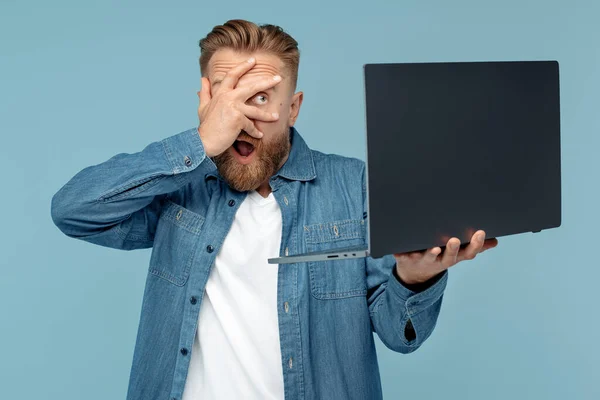 Geschokt baard blond hipster jongeman op zoek naar laptop dekking ogen met de hand over blauwe achtergrond. — Stockfoto