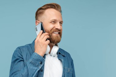Yakışıklı sakallı hippi sarışın genç adam mavi arka planda akıllı telefonla konuşuyor..