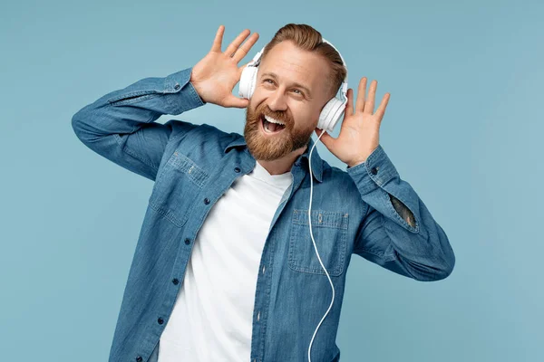 Happy Bearded blonde hipster man in jeans shirt luisteren muziek in wit stijlvolle hoofdtelefoon over blauwe achtergrond. — Stockfoto