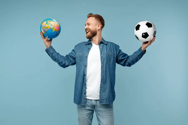 Positieve bebaarde blonde hipster man in medisch masker met klassieke voetbal en wereld bol over blauwe achtergrond. — Stockfoto