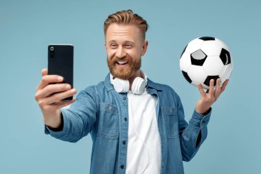 Gülümseyen sakallı hippi sarışın genç adam mavi arka planda klasik futbol topu tutarak akıllı telefondan selfie çekiyor..