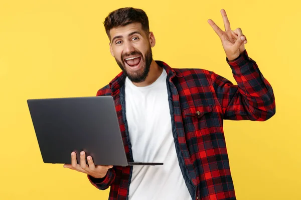 Gelukkig bebaarde hipster jongeman toont vrede gebaar op notebook of computer over gele achtergrond. Open mond. — Stockfoto