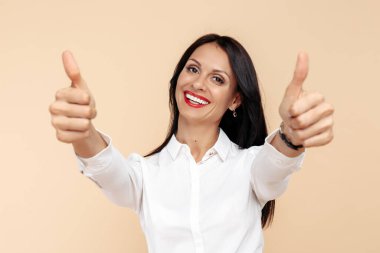 Olumlu genç, modern iş kadını bej arka planda başparmak gösteriyor. Başarı ve kazanan konsepti.