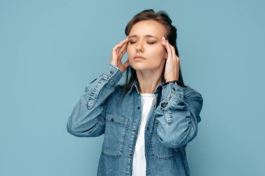 Yorgun genç bir kadın ağır baş ağrısı çekiyor. Şakakları ovuyor, mavi arka planda gözleri kapalı tutuyor..