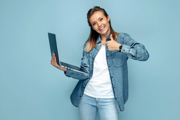 Улыбающаяся молодая женщина в джинсах, указывающая пальцем на компьютер на синем фоне.