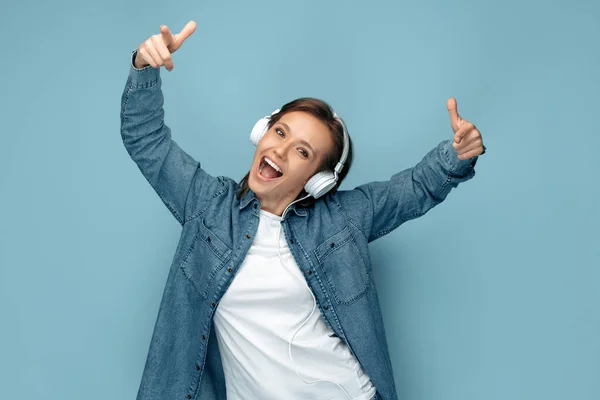 Fröhliche junge Frau in Jeanshemd mit Kopfhörern, die vor blauem Hintergrund tanzt. — Stockfoto