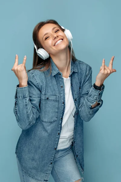 Positive junge Frau im Jeanshemd mit Kopfhörern und Rock-Geste vor blauem Hintergrund. — Stockfoto