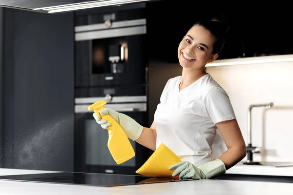 Mujer feliz haciendo las tareas domésticas mientras usa un aerosol en la cocina. Mujer rociando detergente en la placa de inducción. — Foto de Stock