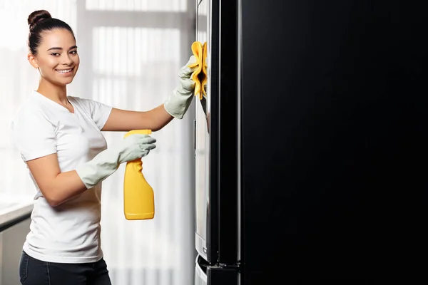 Young woman with cute smile doing the housework while using a spray in the kitchen. Female cleaning refrigerator. — Stock Photo, Image