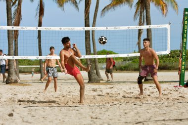 Young Men Kick A Soccer Ball On Beach Volleyball Court clipart