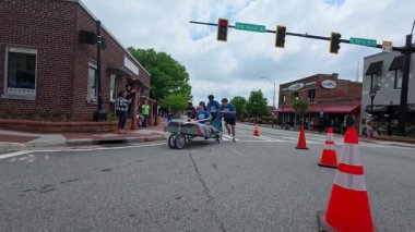 Lawrenceville, GA / USA - 26 Nisan 2025: Yavaş çekim ekibi, 26 Nisan 2025 'te Lawrenceville, GA' da evsizler için yerel bir bağış toplama etkinliğinde, tekerlek üzerinde ev yapımı bir yatağı sokağa iterken görülüyor..