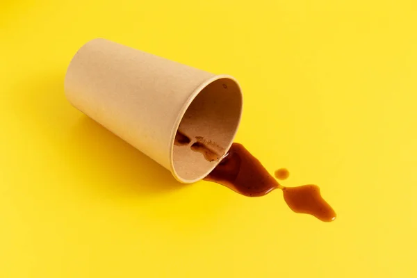 Black coffee spilled from a fallen disposable paper cup on a yellow background.