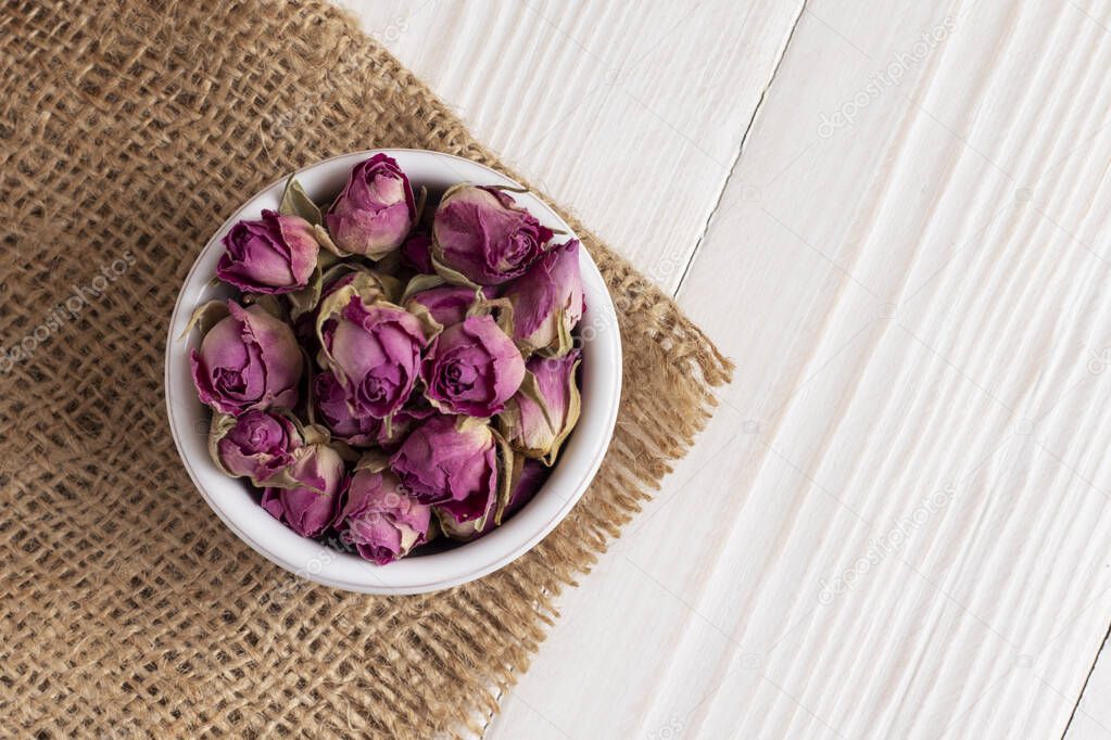 Dried damask rose buds in small bowl on white wooden table. Top view.