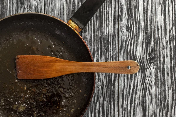 Dirty frying pan with oil and wooden spoon on black wooden table.