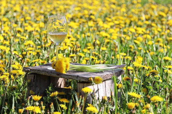 Wine glass with yellow dandelion wine on an old wooden chair in the field.