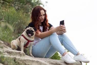Elinde akıllı bir telefonla bir taşın üzerinde oturan kızıl saçlı genç bir kadın. Yanında boksör köpeği oturuyor..