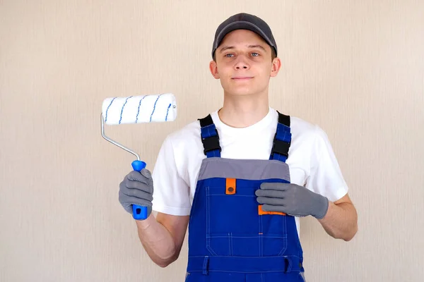 Master painter plasterer in overalls holds a roller in his hand. Blue jumpsuit with shoulder straps. — Stock Photo, Image