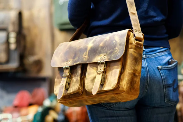 Shoulder travel bag on the shoulder of a young woman in sexy tight ass jeans against the background of other bags of the store. The concept of natural leather products. — ストック写真