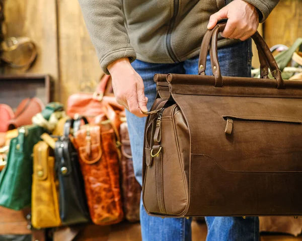 The hand of a man holding a brown travel bag against the background of other bags in the store. The concept of natural leather products. — Stock Photo, Image