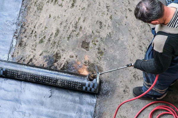 Ein Arbeiter installiert Harzfolie auf dem Dach eines Gebäudes. Wasserdichtes System mit Gas und Feuerfackel. Abdichtung des Daches. — Stockfoto