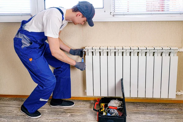 A plumber in a blue jumpsuit is fixing a radiator. With an adjustable wrench, he tightens the tap. A briefcase with a tool at hand. — Stock Photo, Image