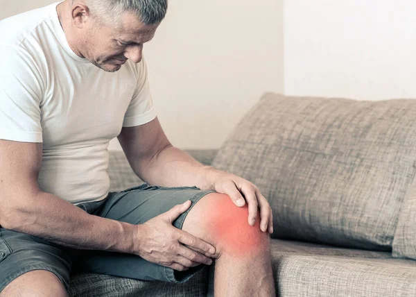 Artritida je onemocnění kloubů. Muž na gauči, mačkající si koleno před nesnesitelnou bolestí. — Stock fotografie