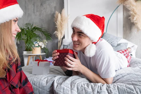 People, gifts and holidays concept. Caucasian couple at home. Young smiling man lying on bed holding gift box and looking at happy woman sitting on the floor next to him, wearing santa hats