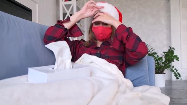 休日の概念 若い女性は病気の兆候を経験しています 喉の痛み 女性は保護医療マスクとサンタの帽子をかぶったリビングルームのソファに横たわっています — ストック動画