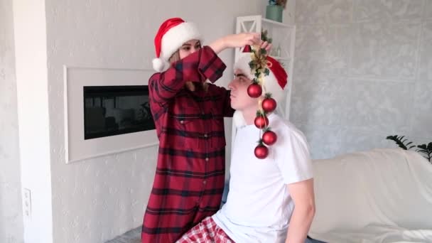クリスマスツリーの代わりにクリスマスボールで男を飾る回避若い遊び心のある女性 サンタの帽子と赤いパジャマを着たカップル スローモーション — ストック動画