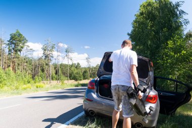 Adam yaz güneşli bir günde yol boyunca park etmiş bir arabanın bagajından bir kaykay çıkardı.