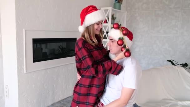 Fiatal játékos nő bolondozik díszítő férfi karácsonyi labdák helyett karácsonyfa. Télapó kalapot és piros pizsamát viselő pár — Stock videók